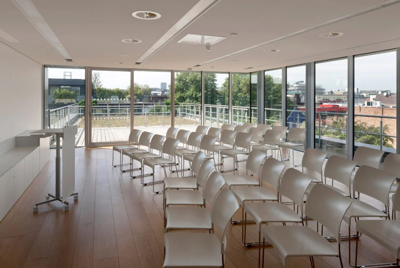 Empty conference room with white chairs and large windows showing an outdoor balcony and cityscape.