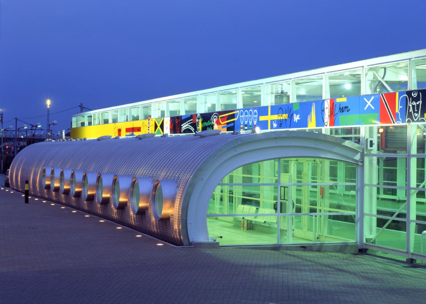 The colourful Tottenham Hale Station entrance with glass doors and a metallic, curved structure, illuminated with greenish lighting, and bright abstract mural that runs along the upper level, during dusk against a dark navy-blue skyline.