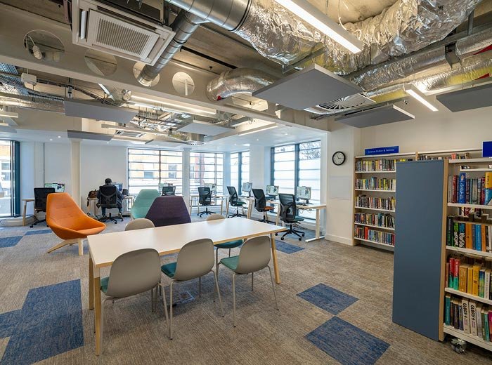 Inside the Northcote Library building with large windows, open ceiling with exposed ductwork, and various chairs including office chairs at desks and lounge chairs, some near a wall of bookshelves.