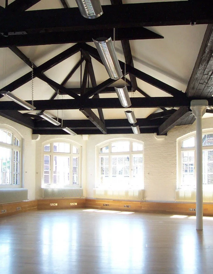 Empty room within the Thomas Neal's Centre wtih large windows, hardwood floor, white brick walls, and a high ceiling with black wooden beams and hanging fluorescent lights.