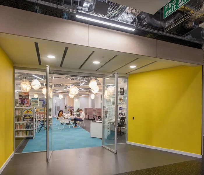 View of a modern library or community space with yellow walls, glass doors, hanging lights, bookshelves, and people reading and working inside.