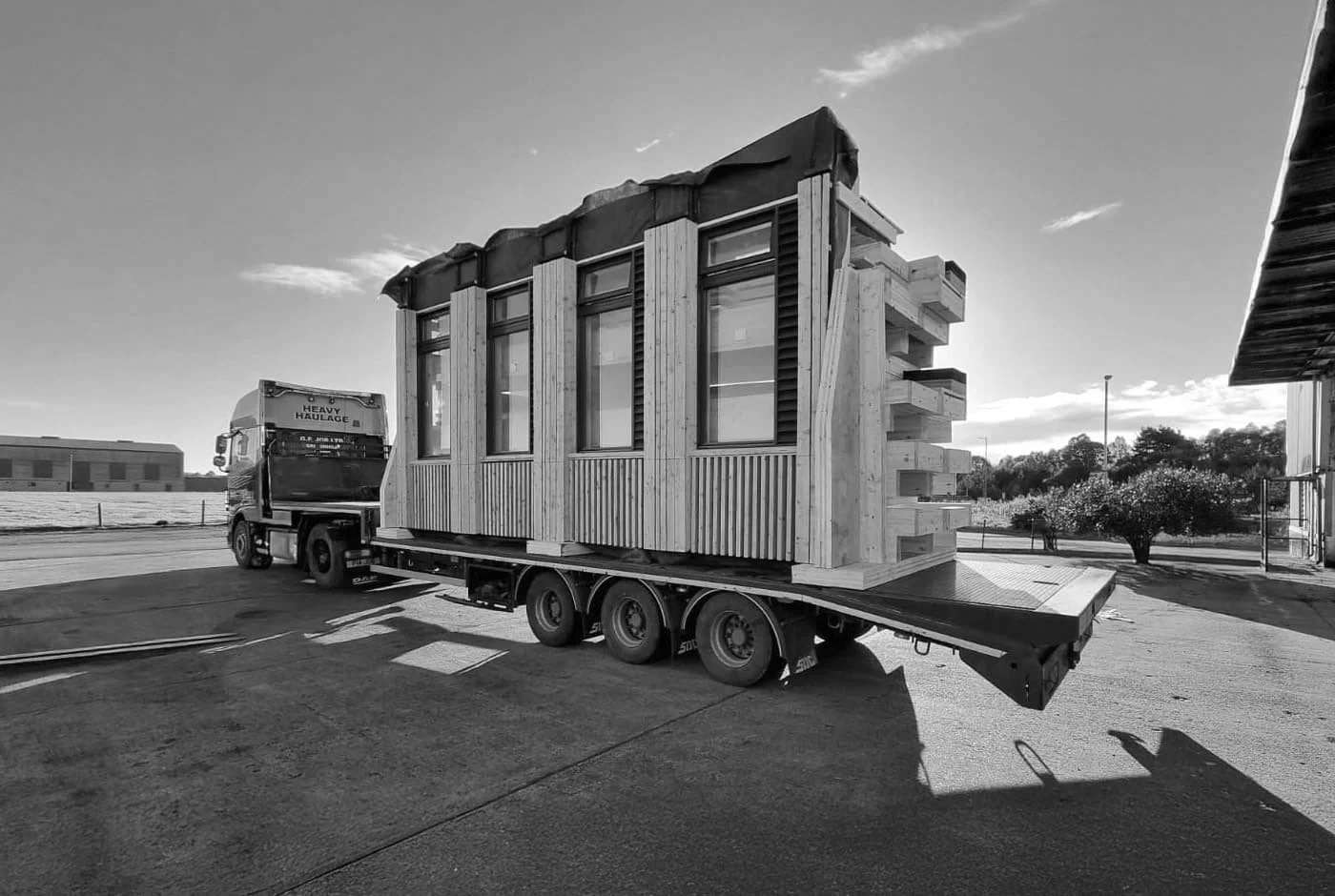 A flatbed truck transporting a small, modern house made of wood, with large windows and a covered roof, parked on a paved area under a clear sky.
