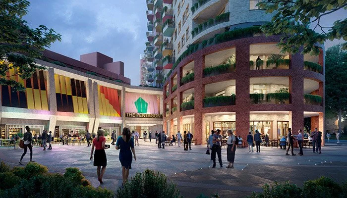 People walking and socializing in an urban plaza at dusk, with a modern multi-story building and a colorful, illuminated structure in the background.