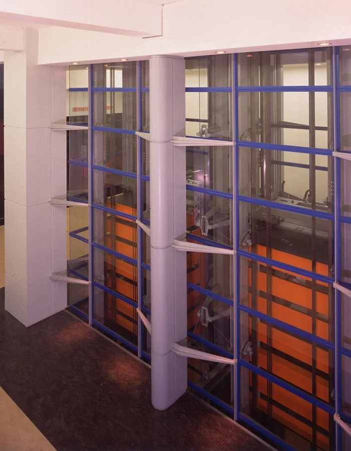 An elevated side-view of the internal workings of the Science Museum elevator with visible internal mechanisms through the glass walls, displaying orange panels against the blue frame panels, with grey support beams outside.