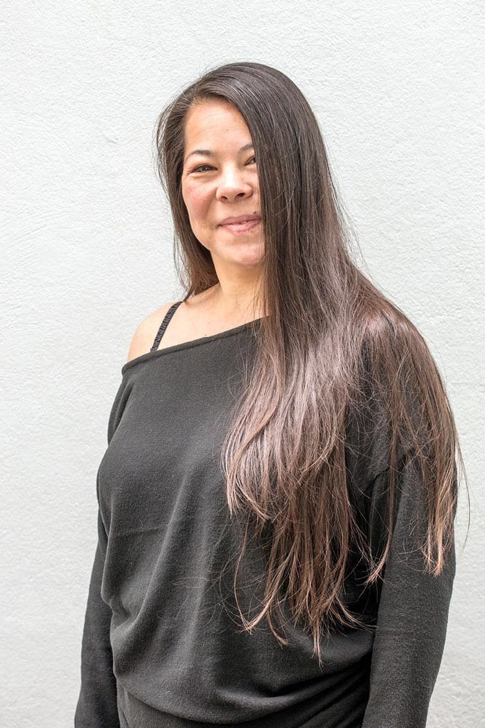 A woman with long brown hair wearing a black off-shoulder top, standing against a plain white wall.