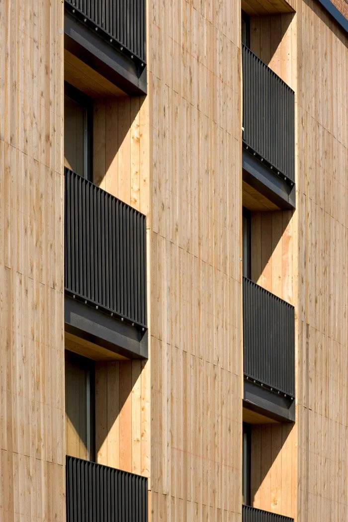 Close-up of a building facade with light wood siding and black metal balcony railings, with shadowed recessed balconies.