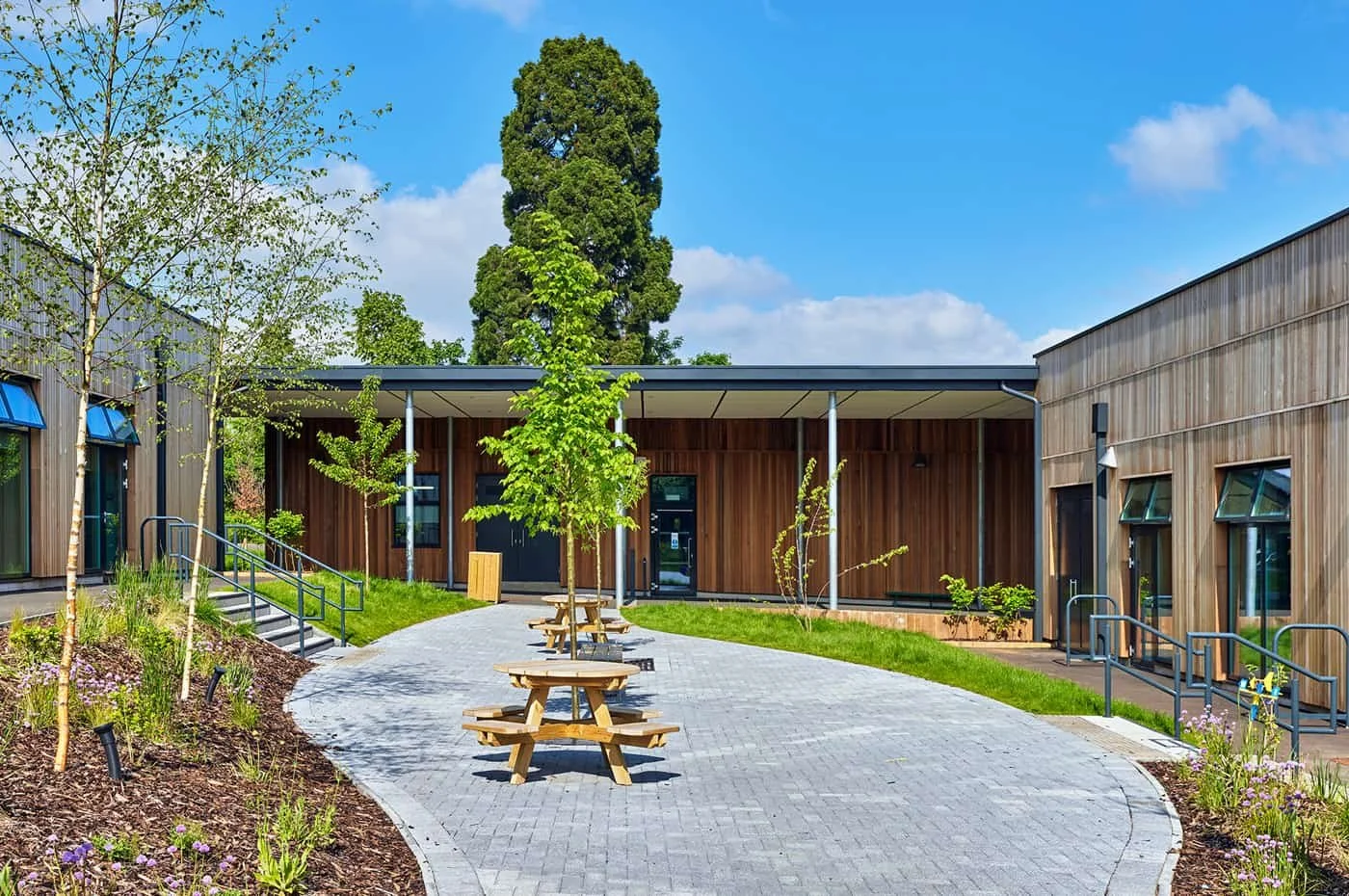 Modern building courtyard with landscaped garden, trees, picnic tables, and a paved walkway under a partly cloudy sky.
