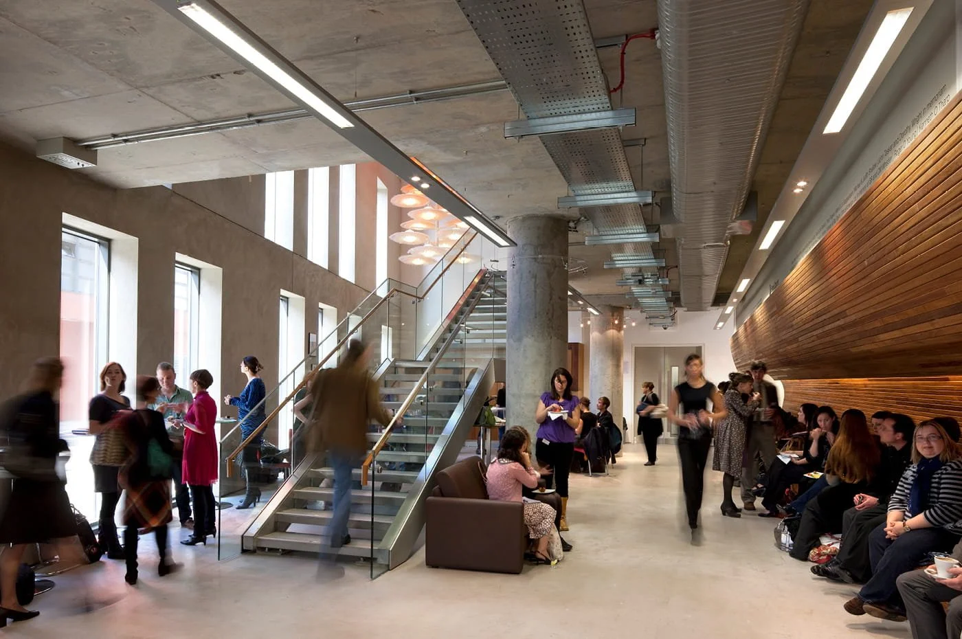 Interior of a modern, busy entrance area with people sitting, standing, and walking, featuring large windows, a staircase, and wood-panelled walls.