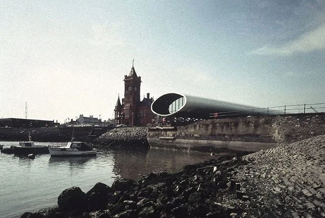 A waterfront scene at Cardiff Bay, with boats, a historic red brick building with a clock tower, and a modern metallic tunnel structure of Inner Harbour over a walkway.