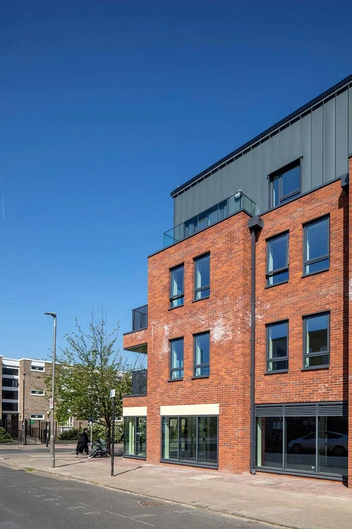 A modern red brick building with large ground-floor windows and smaller upper windows, with black accents, near a sidewalk with a tree and pedestrians, under a clear blue sky.