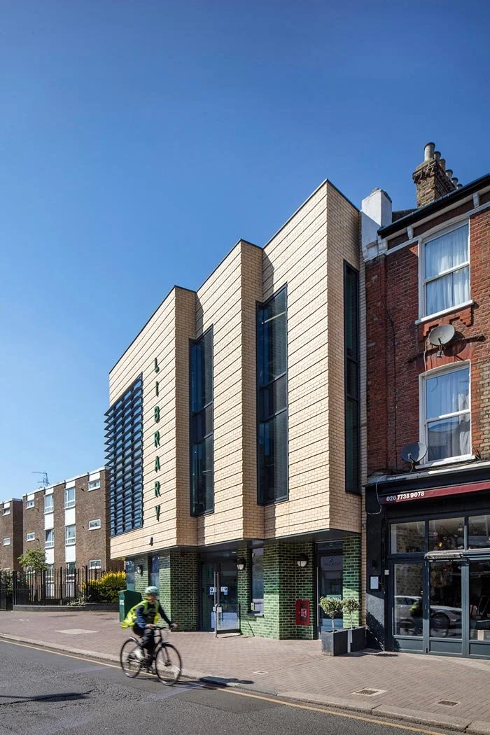 The upper section of beige brick Northcote Library and green glazed brick on ground floor, with tall narrow windows on a city street, adjacent to an older red brick building with satellite dishes, under a clear blue sky, with a cyclist riding past.