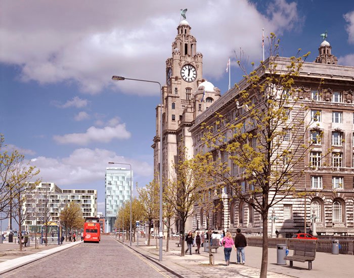 Liverpool waterfront with historic Royal Liver Building and pedestrians on the sidewalk, with the light grey tall structure of the  New World Square multi-story modern building in the distance.