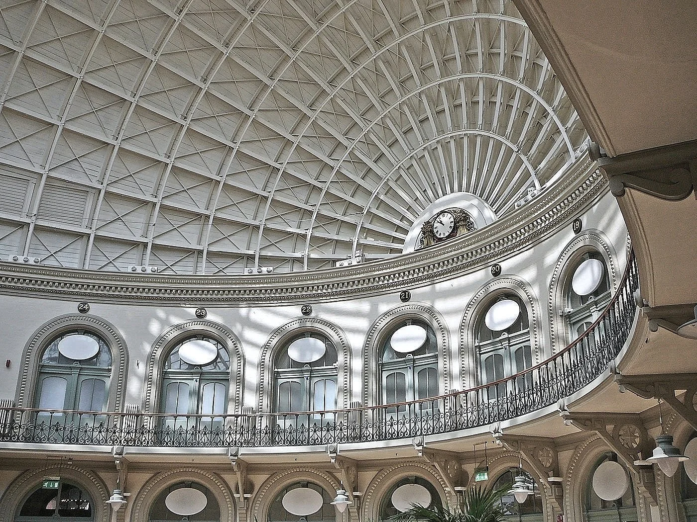 Interior closeup of the Grade 1 listed Corn Exchange in Leeds historic interior building with a curved balcony, ornate trim, large arched windows with oval openings, a decorative clock, and a glass domed ceiling with metal framework.