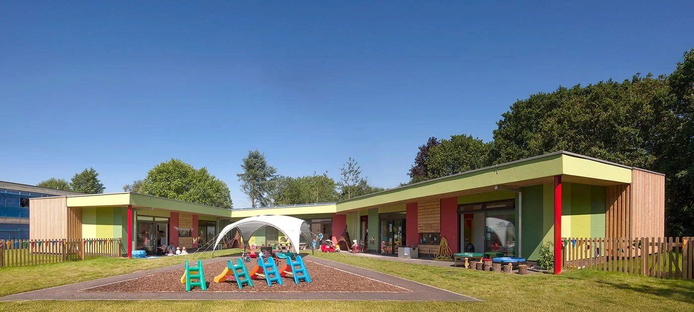 A panoramic view of the Enchanted Wood Nursery from the inner play area, colourful modern preschool building with a playground in front, including slides, climbing structures, and benches, surrounded by a grassy yard and trees under a blue sky.