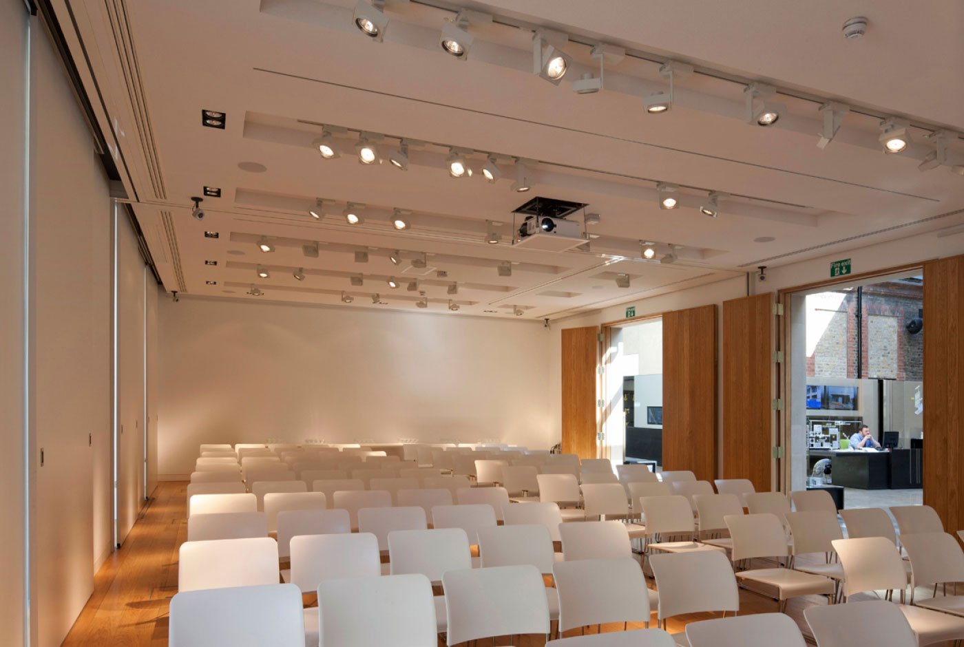Empty conference room with white chairs arranged in rows, large windows with natural light, and a projector on the ceiling.