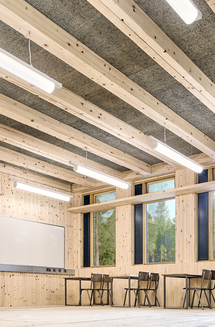 Classroom with wooden walls, ceiling beams, large windows, whiteboard, and chairs and desks.