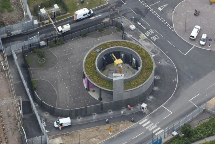 A birds-eye view of the Pudding Mill Pumping Station completed site with it's green-living circular roof, surrounded by a security fence, located in an urban area near roads and a few parked vehicles.
