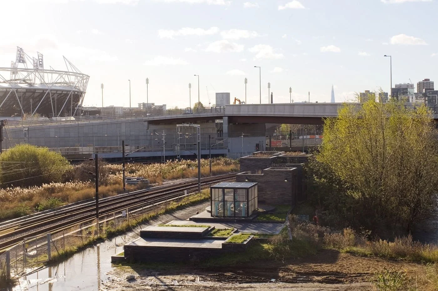 Cityscape with railway tracks, a glass greenhouse, green trees, a bridge in the background, and a modern stadium to the left, under a blue sky with scattered clouds, with the Stratford Box Pumping Station in the near foreground.