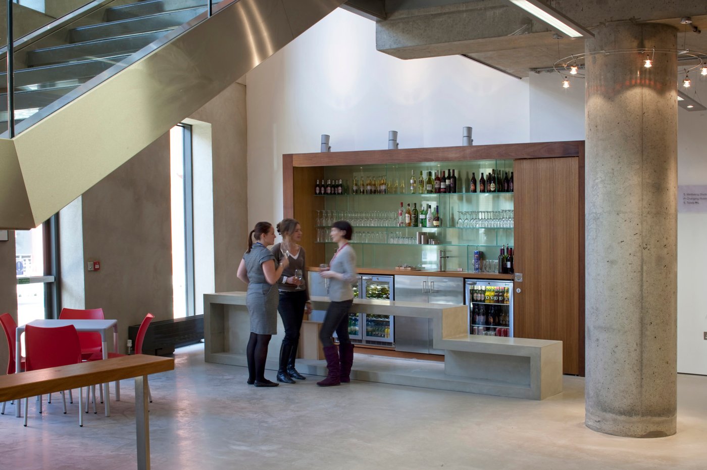 Three women stand and converse at a modern bar area inside a building. The bar has glass shelves with bottles and glassware, and a small refrigerator under the counter. Red chairs and a table are to the left, along with a large concrete column and staircase above.