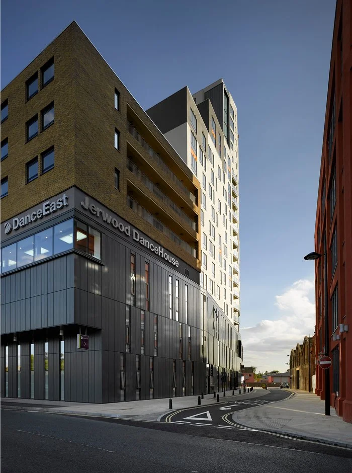 Modern multi-story building with signage for Jerwood Dance House, situated on a street corner with a curved road, adjacent to traditional brick buildings, under a clear blue sky.
