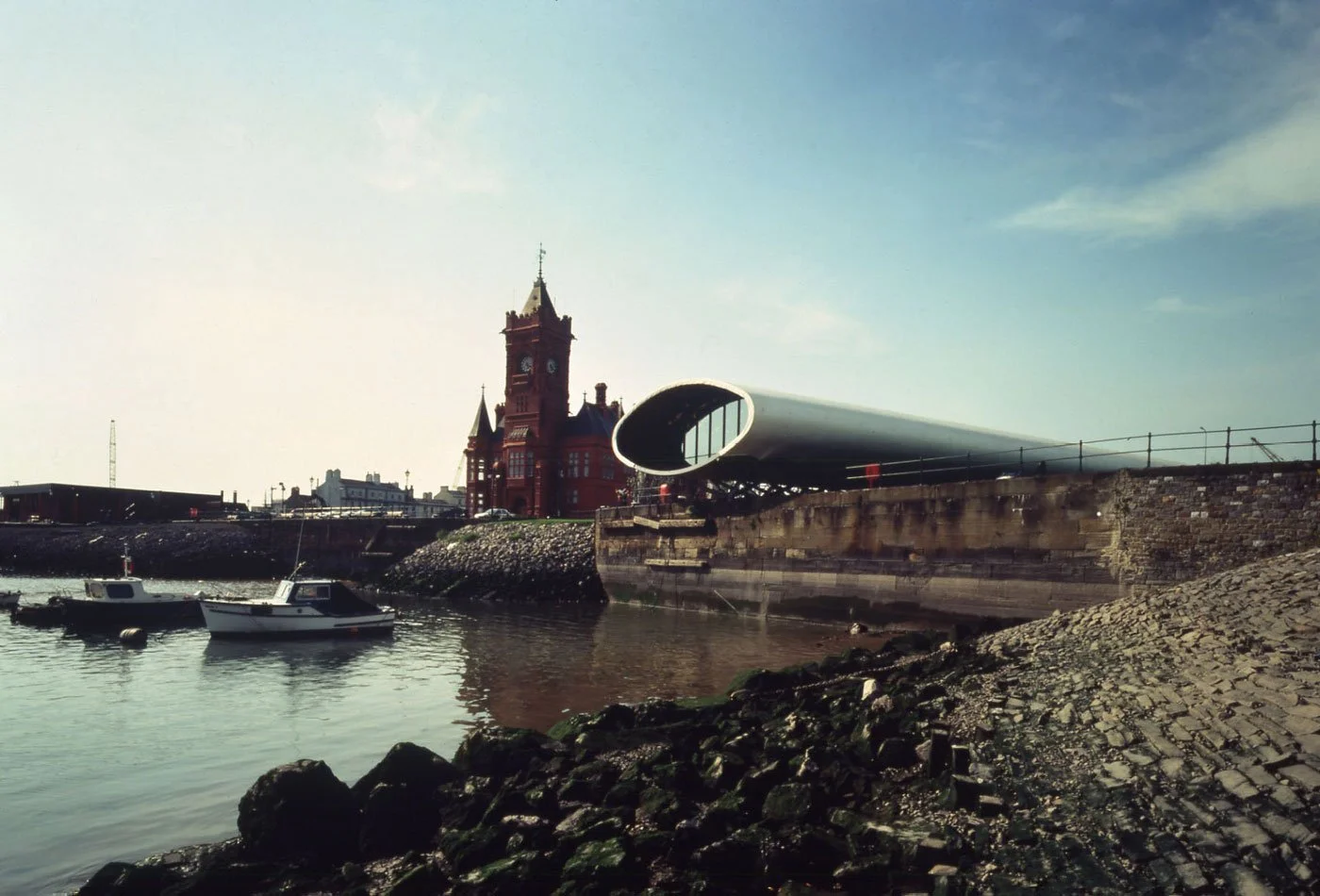 The view of Cardiff Bay Inner Harbour with small boats and a historic red brick building with a clock tower in the background, the 70 metre long squashed tube of elliptical steel rings that is the Visitor Centre extends over the water.
