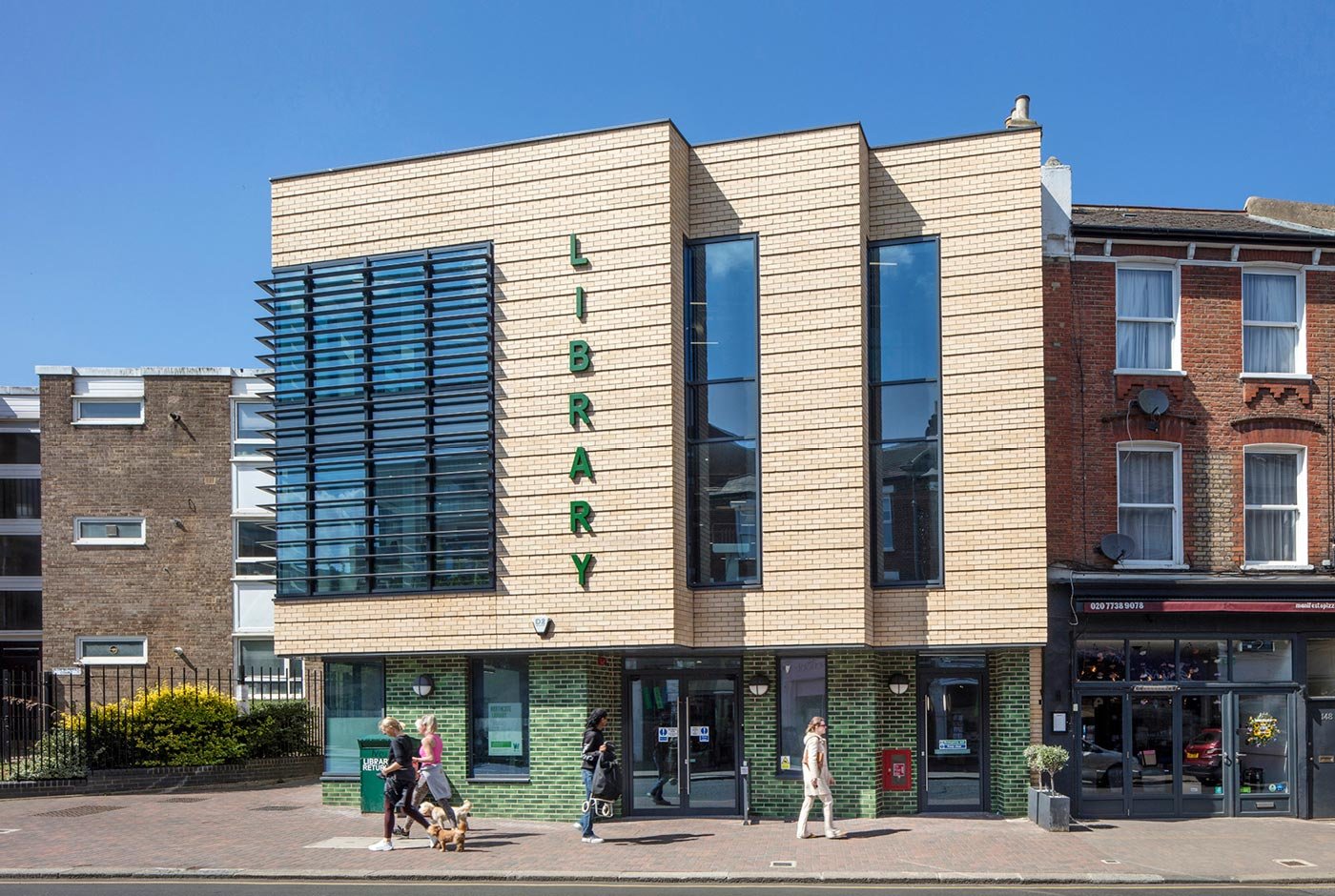 A side view of the Northcote Library on the Wandsworth high street with pedestrians walking past in either direction with the  green glazed brickwork behind them on a bright summers day.