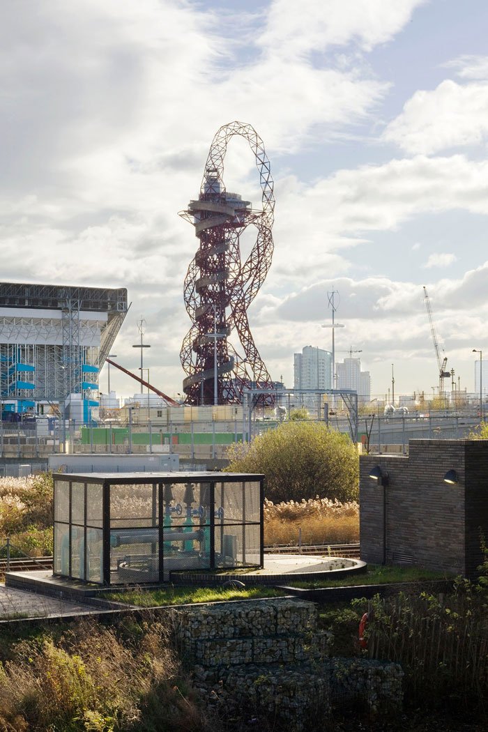 The Olympic ArcelorMittal Orbit structure and various other buildings and cranes in the background, and a small cage-enclosed shelter of one of the Stratford Box Pumping Station building the foreground.
