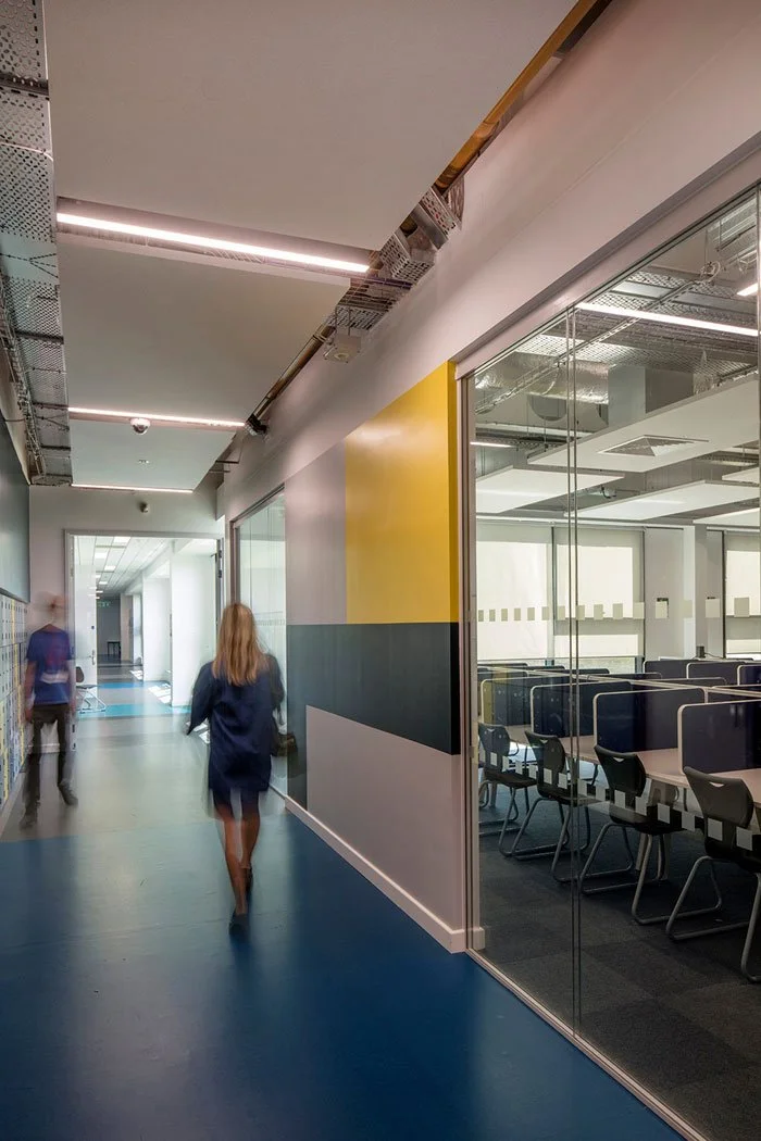 People walking down a corridor in an office building, with one person in a navy blazer and a woman with long blonde hair. To the right, there is a glass-walled conference room with chairs and tables inside. The ceiling has exposed pipes and lighting.
