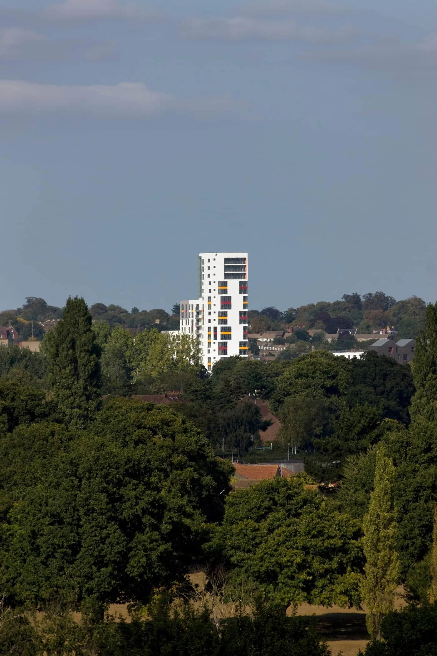 A modern white high-rise building with colored window accents, surrounded by green trees, under a partly cloudy sky.
