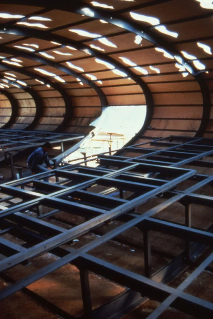 Under construction interior shot of the the 70 metre long squashed tube of the Visitor Centre, a large curved wooden structure with metal framework and beams, with a worker welding or assembling metal parts inside.