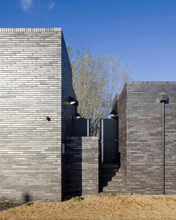 A side elevation close up of the Stratford Box Pumping Station's modern building with neat square lines with black doors, exterior stairs, and outdoor lighting, under a clear blue sky with trees in the background.