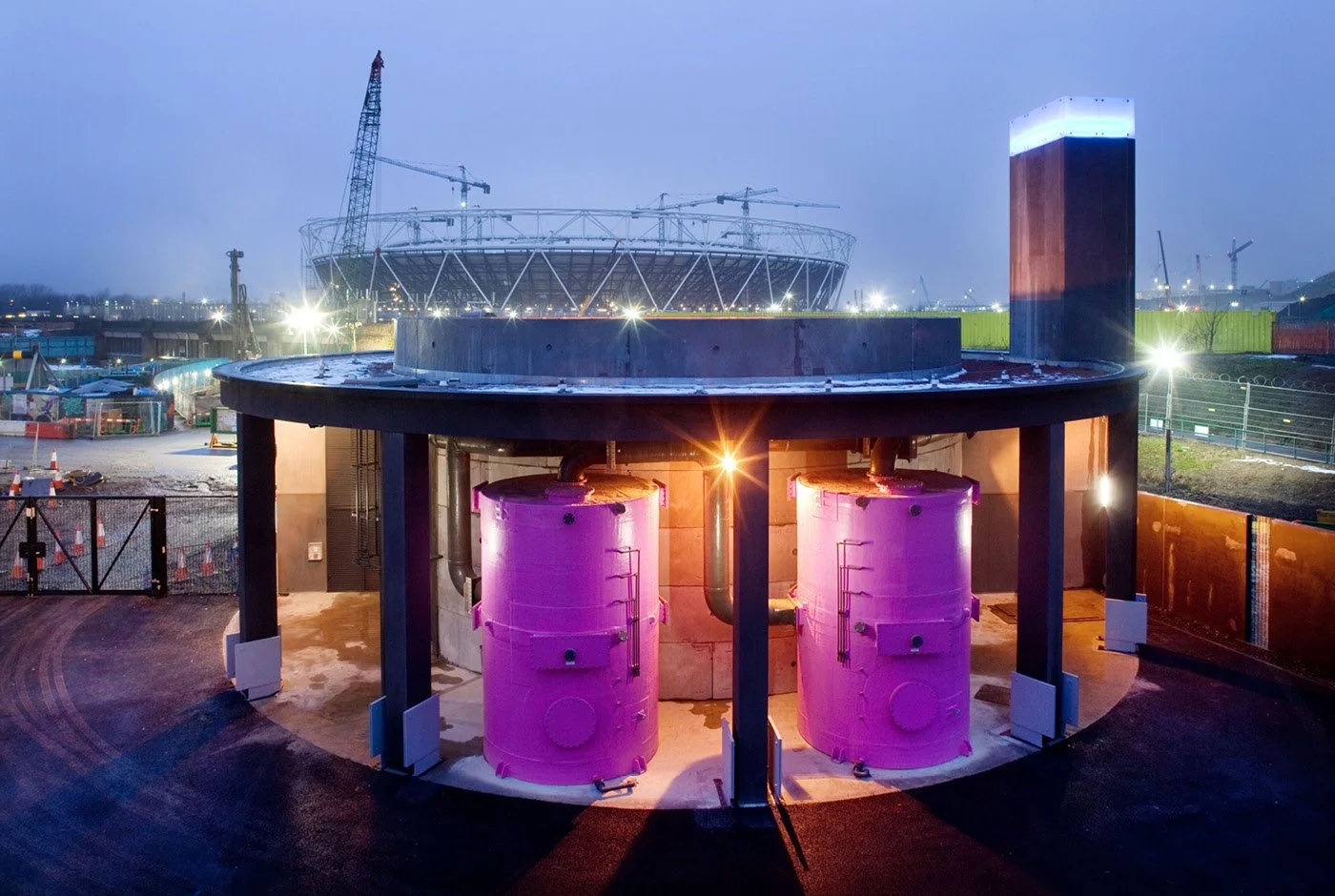 Cityscape at dusk showing industrial structures of the Pudding Mill Pumping Station, with large pink storage tanks illuminated, and it's modern tower glowing blue lantern at the top.