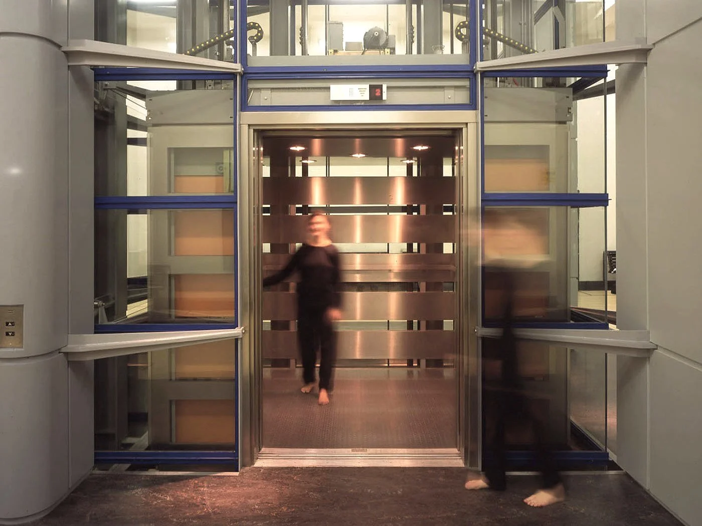 People entering/exiting the main elevator at the ground floor of Science Museum, London, one person appears blurred with only their legs visible, the other is inside the elevator, background shows modern interior design.