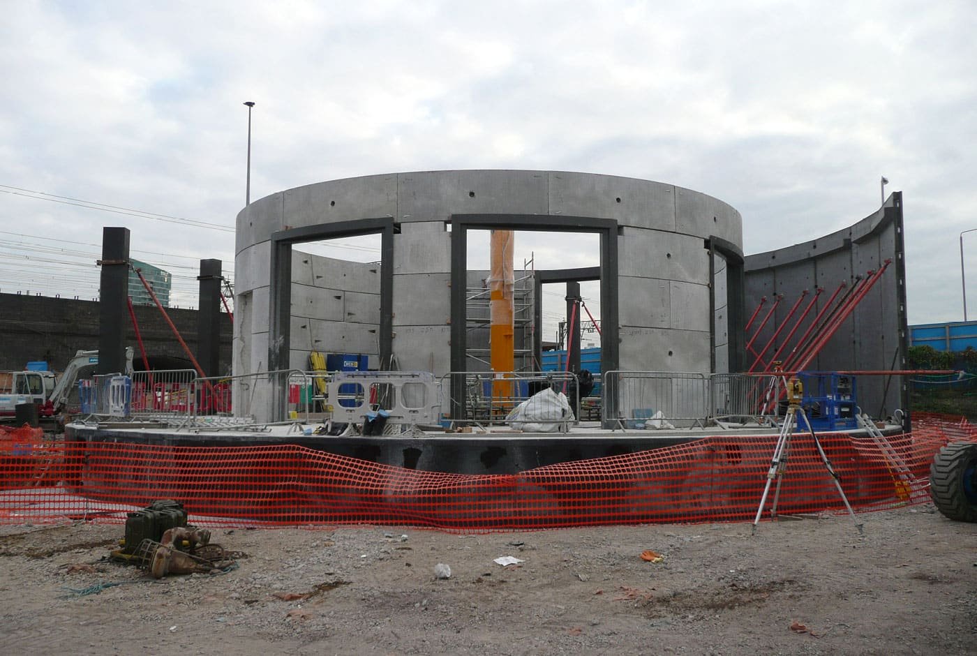 Construction site with a circular concrete structure in progress, surrounded by safety barriers and construction equipment under a cloudy sky.