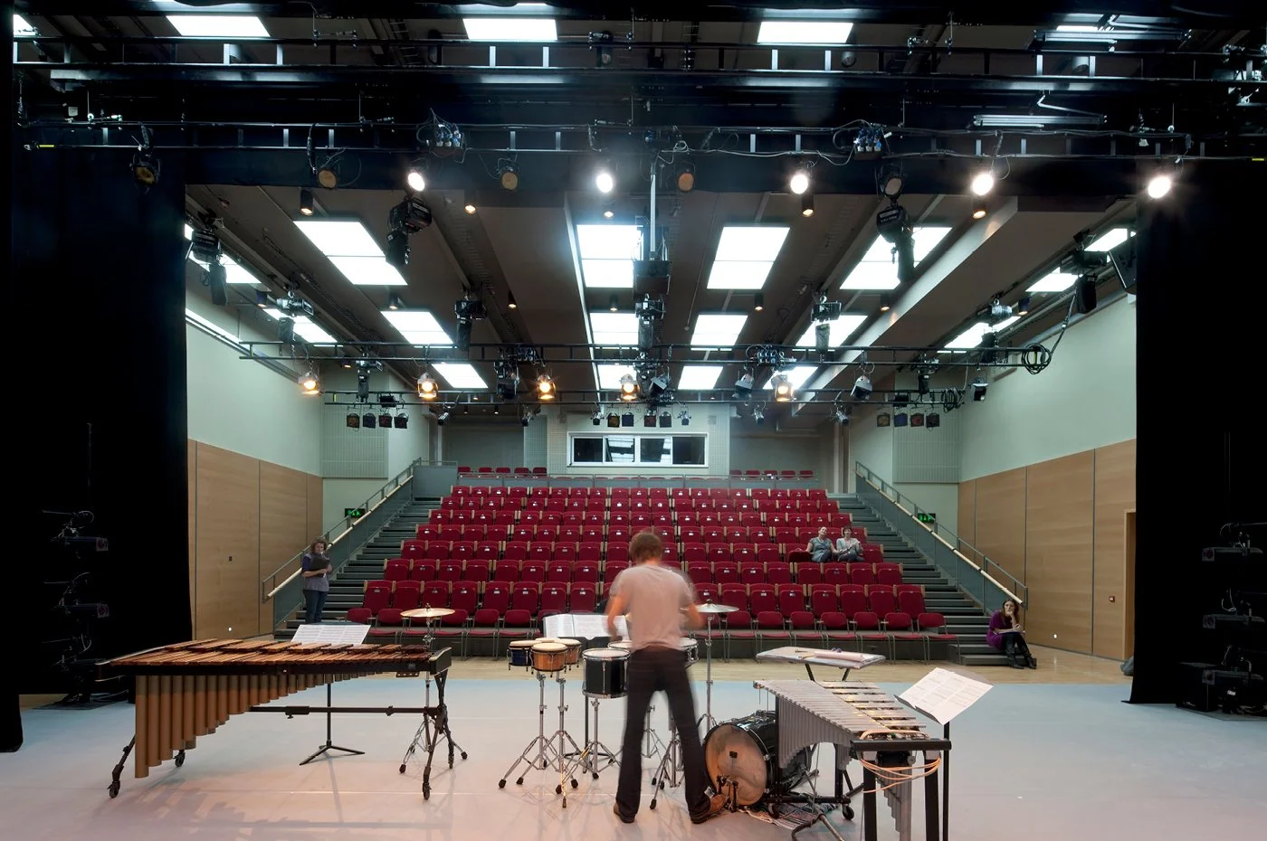 An empty theater or auditorium with a stage set up for a musical or performance. Instruments, including drums, xylophone, and percussion, are on the stage. Airplane interior with red seats and a staircase, and a few people seated and standing in the audience area.