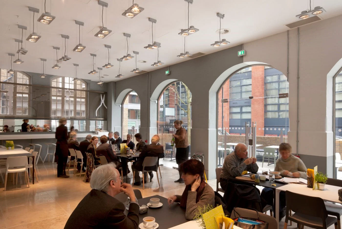 People dining in a modern restaurant with large arched windows, pendant lighting, and a mix of seated patrons and staff serving customers.