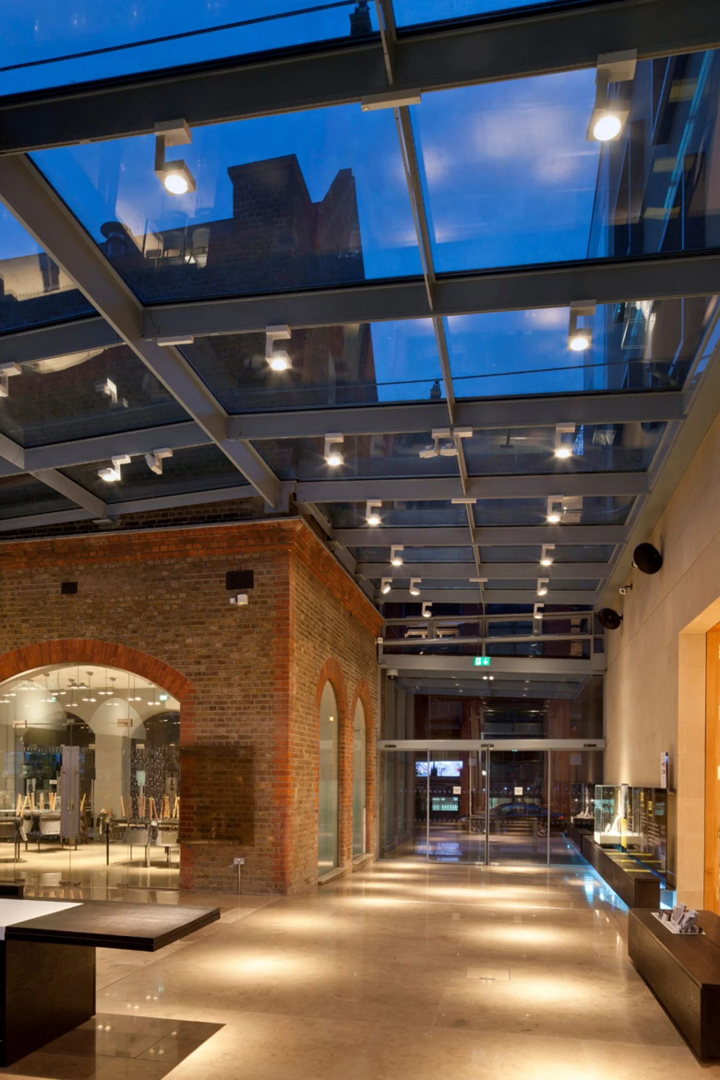 Indoor shopping mall with brick walls, glass windows, and a glass ceiling. Bright ceiling lights illuminate the space, and storefronts are visible along the corridor.