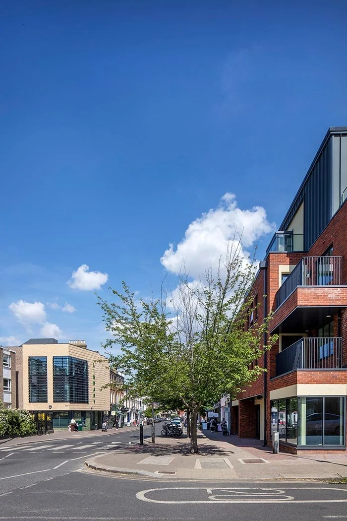 City street scene in Wandsworth with a tree in the middle of a pedestrian island, with the modern Housing buildings on the right and the Northcote Library in the distance on the left against a blue sky with light white clouds.