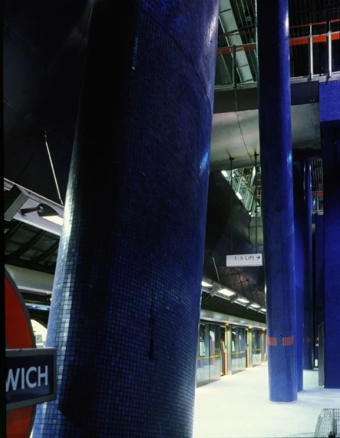 An extreme closeup of one of North Greenwich underground station large blue cylindrical columns, a train on the platform, and signs indicating the lift and other directions.