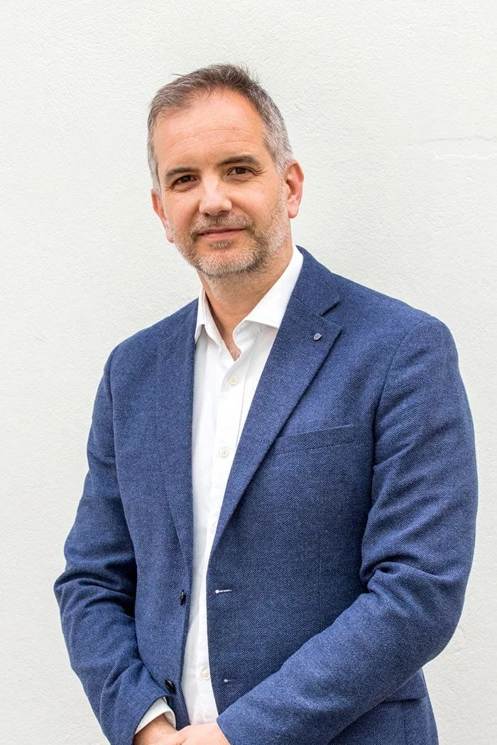 A middle-aged man with gray hair and a beard, wearing a blue blazer and white shirt, standing against a white wall.