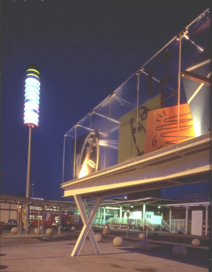 A nighttime outdoor view of Tottenham Hale Station with it's X-shaped pillars holding up the protruding first floor, displaying bright artwork behind class walls, a tall pole with bight illuminated lighting is in the background.