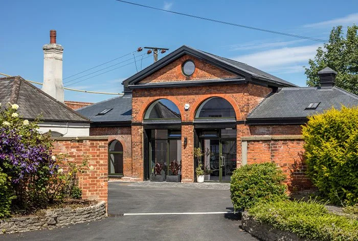 A red brick building with large arched windows and a black roof, surrounded by greenery and shrubbery.