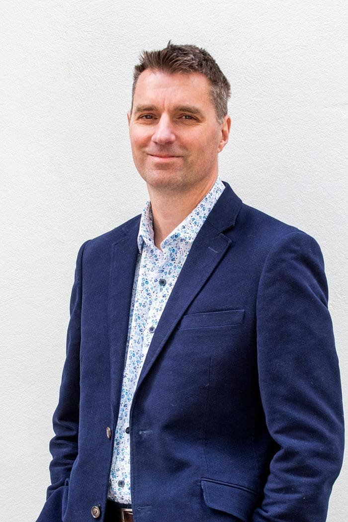 A man in a dark blue blazer and white patterned shirt standing in front of a plain white wall.