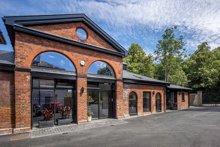 A view looking at an angle to the front of the main Mascalls Stable Block brick building with large arched windows and a black roof, surrounded by trees, with a parking lot in front.