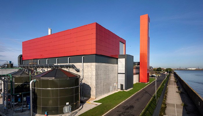 A modern industrial building with a red upper section, gray lower section, and black piping, located beside a waterway with a pathway and railing.