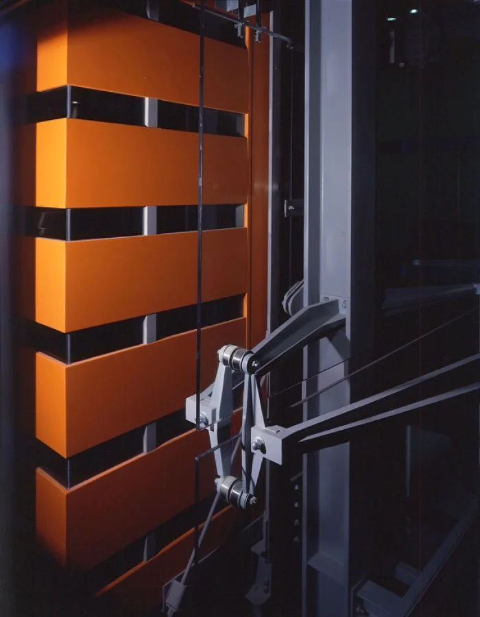 A close-up of an distinct orange panels with sleek lines of the Science Museum elevator area with the mechanical system inside and metal components and cables on display.