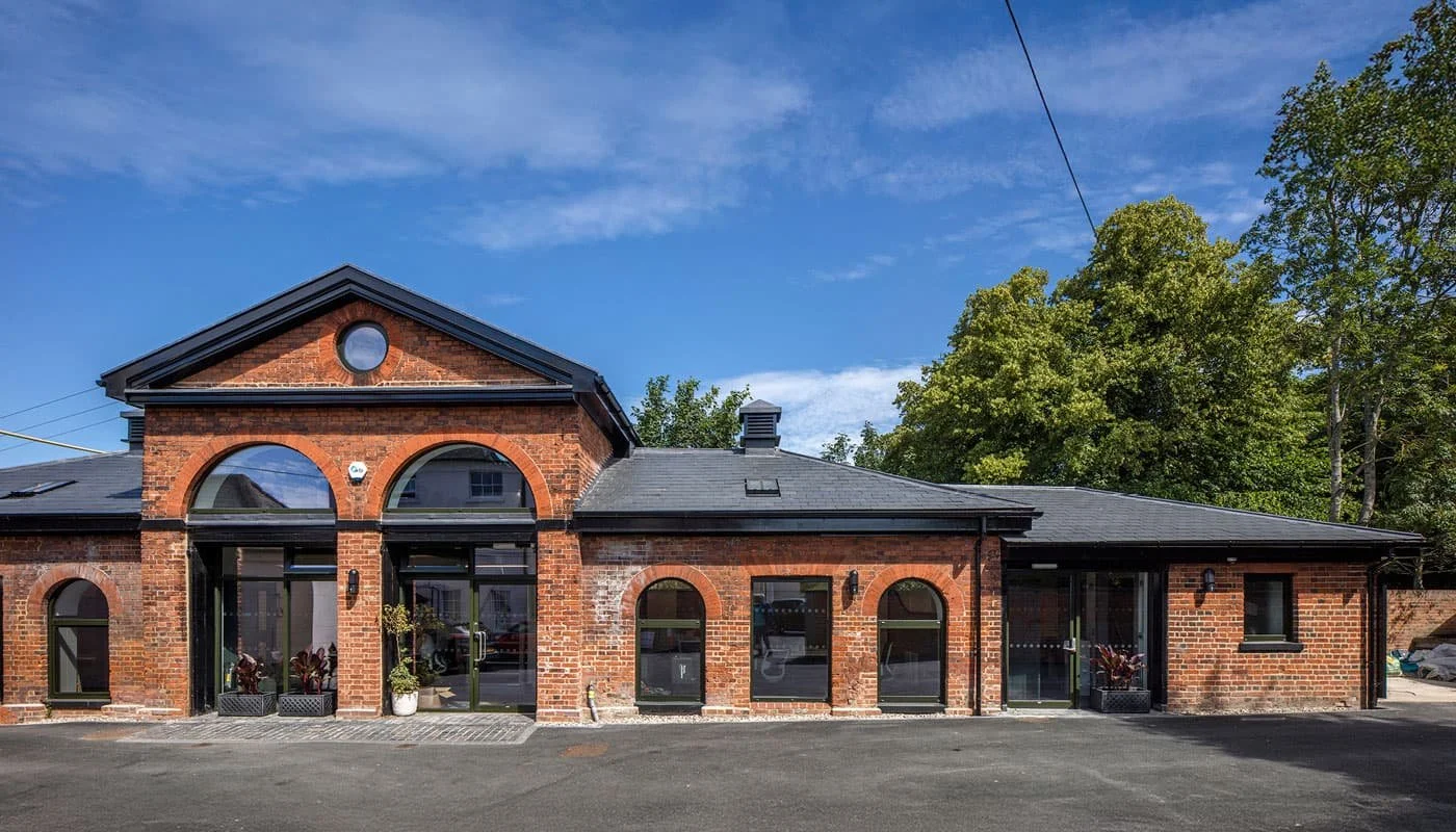 Outside the main entrance to Mascalls Stable Block with large arched windows and doors, set against a backdrop of trees and a blue sky with some clouds.