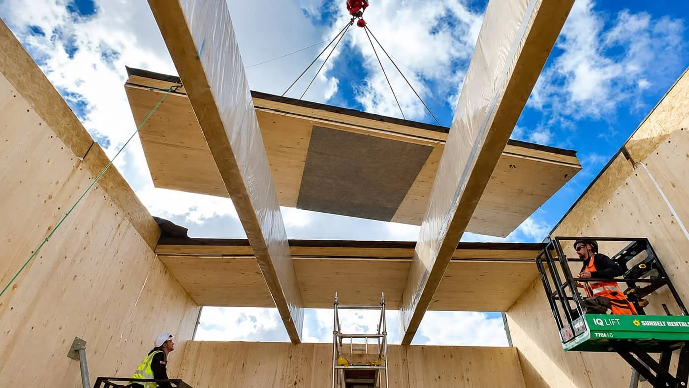 Construction workers and equipment lift large, prefabricated wooden wall and roof panels into place for building a structure against a blue sky with clouds.