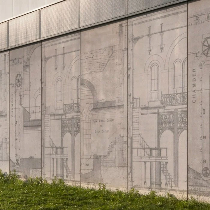 A close up of one side area of the Crossness Sludge Cake Building showing the engraved concrete panels, embossed with historic engineering drawings from the nearby Abbey Mills Pumping Station, with a small amount of grass where they meet the ground.