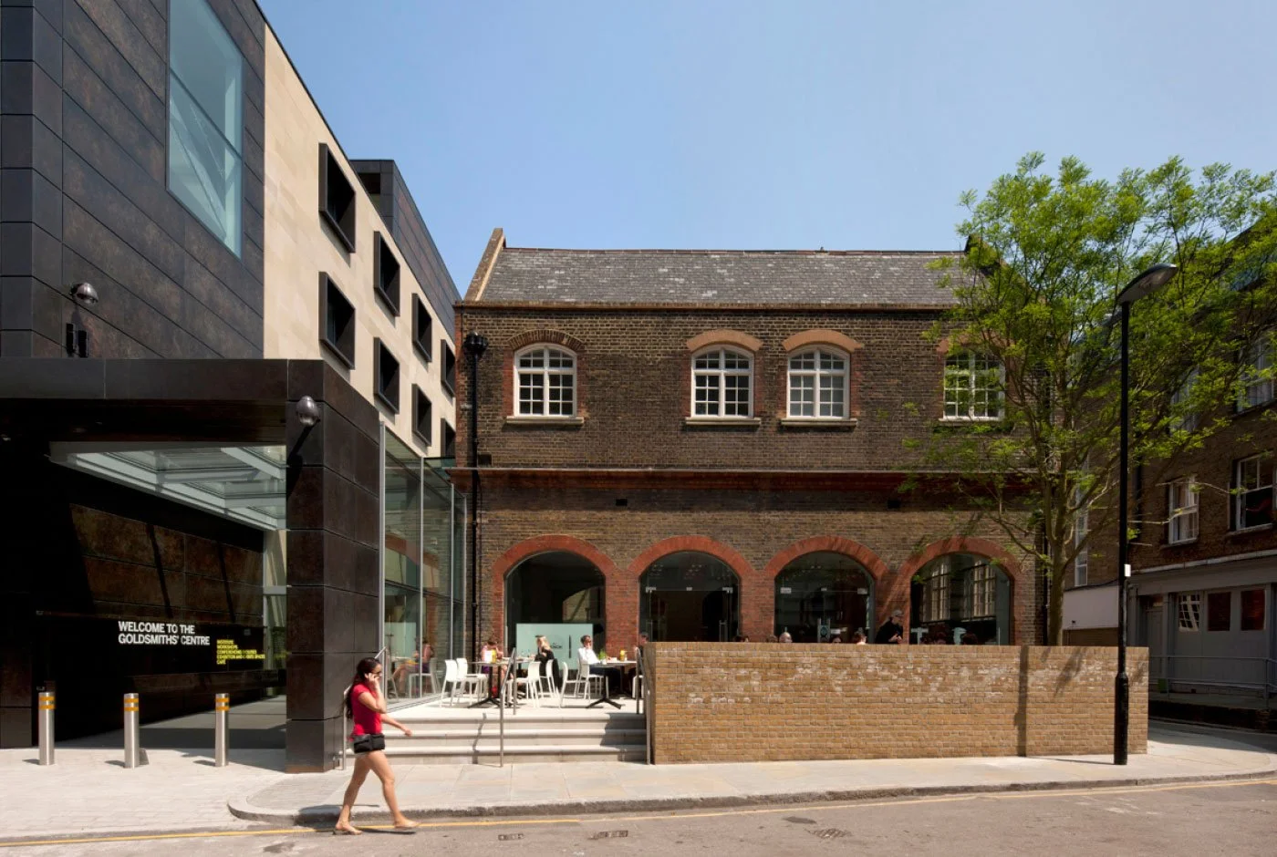 The entrance to Goldsmiths' Centre, with dark archway, showing a mix of the Grade II listed Victorian school with arched windows together with new materials, with people sitting outside a café and a woman walking on the sidewalk, under a blue sky.